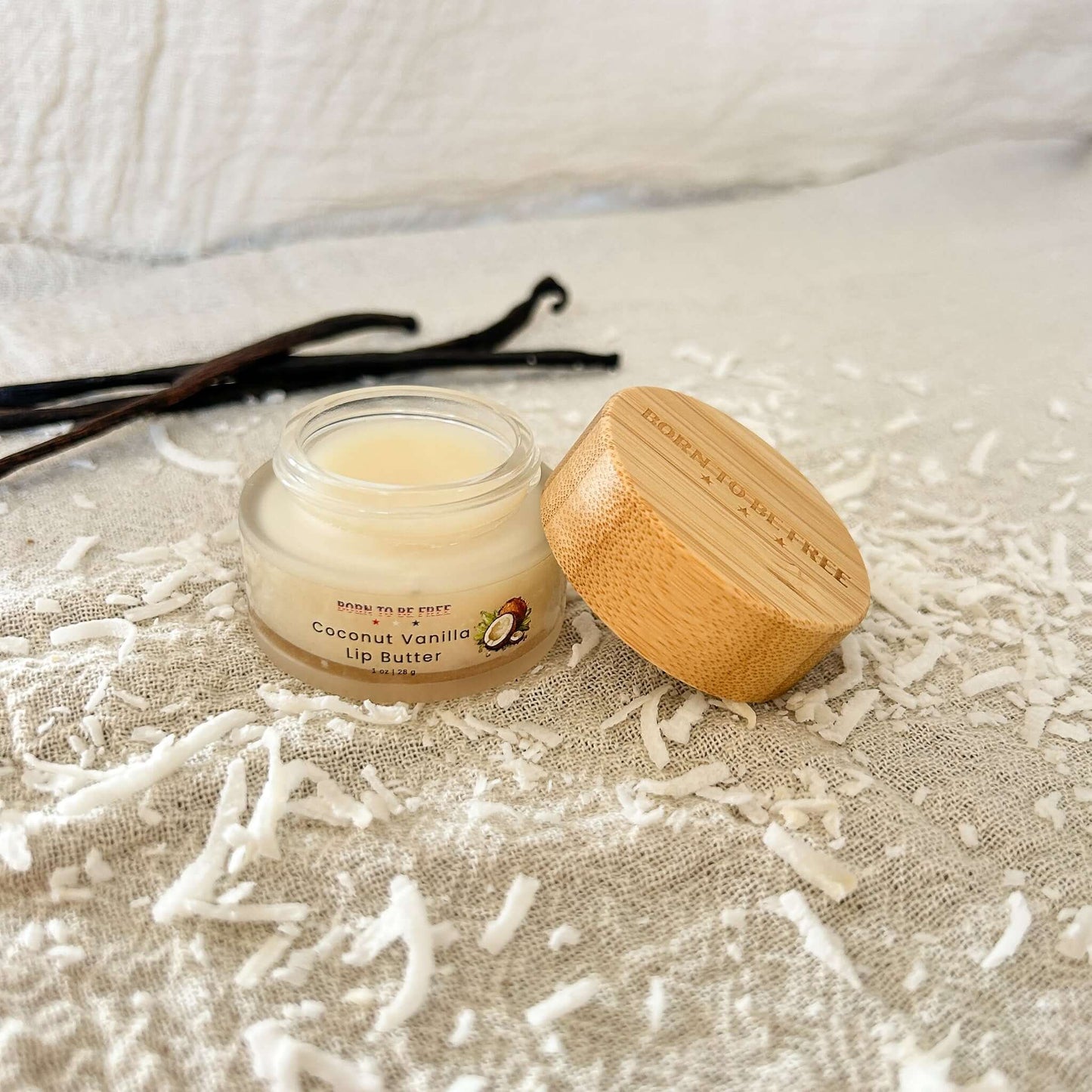 Coconut Vanilla Lip Butter in a glass jar with bamboo lid, surrounded by coconut shavings and vanilla sticks.