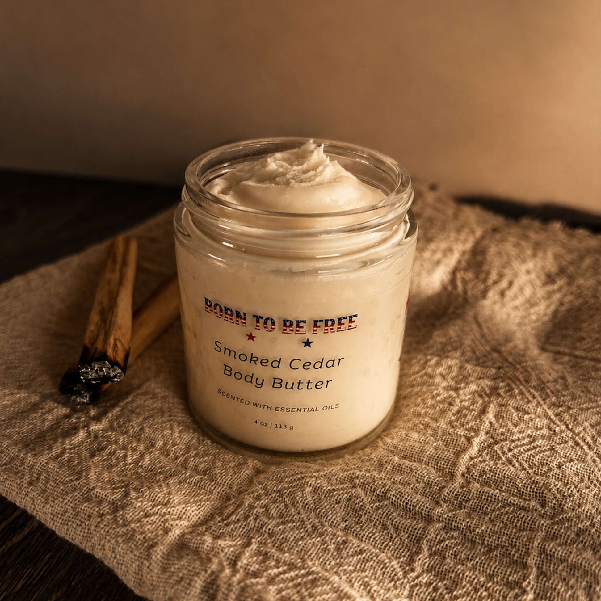 Smoked Cedar Body Butter in a glass jar on a textured cloth with cinnamon sticks.