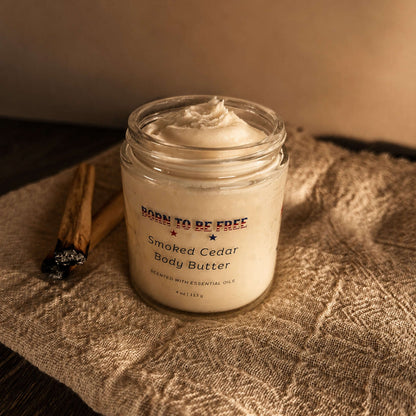 Smoked Cedar Body Butter in a glass jar on a textured cloth with cinnamon sticks.