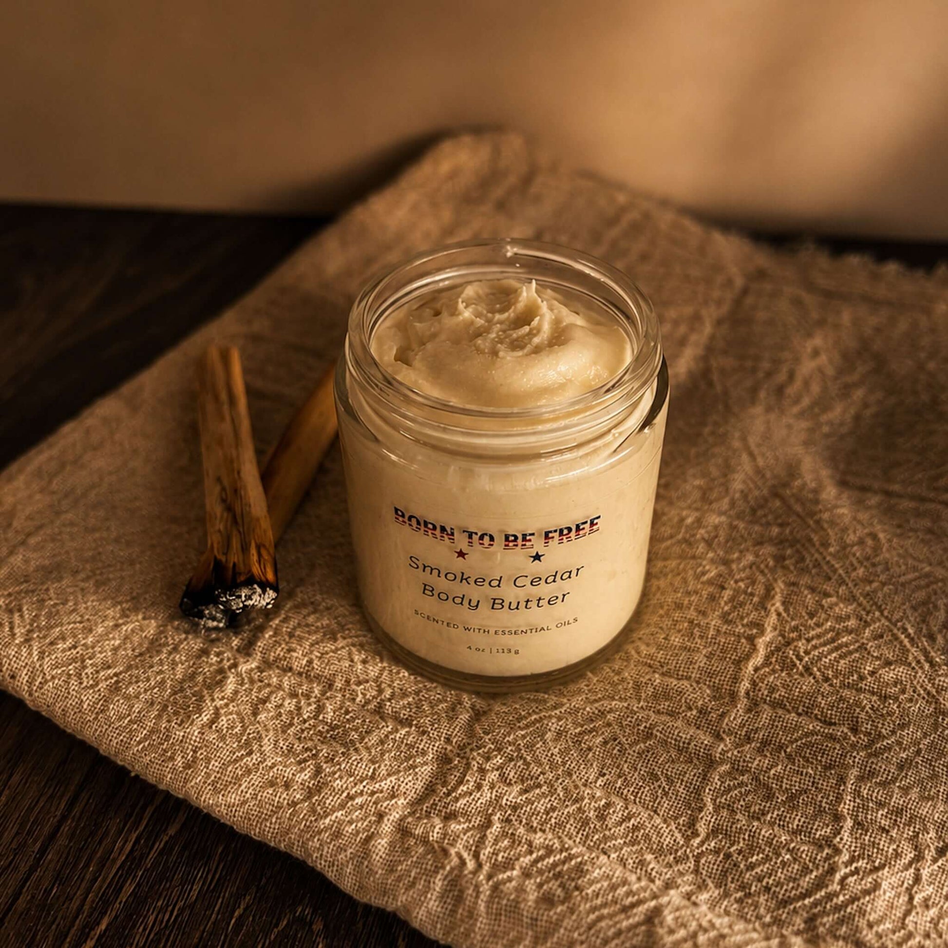 Smoked Cedar Body Butter in a jar, surrounded by cedar sticks, showcasing a rich, whipped moisturizer.