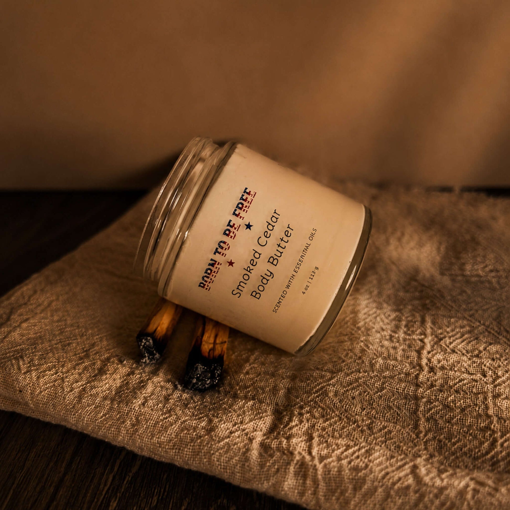 Smoked Cedar Body Butter jar on a textured cloth with burnt wood accents, showcasing a natural skincare product.