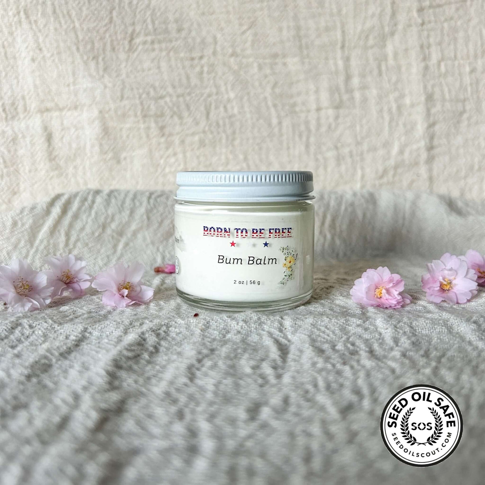 Bum Balm - Baby Diaper Cream with Tallow + Zinc in a glass jar surrounded by pink flowers on a textured background.