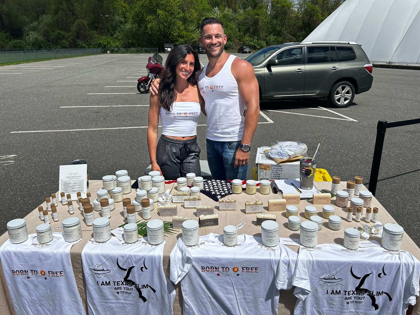 Two vendors at a market showcasing Feeding Forward T-shirts and various products on a table.