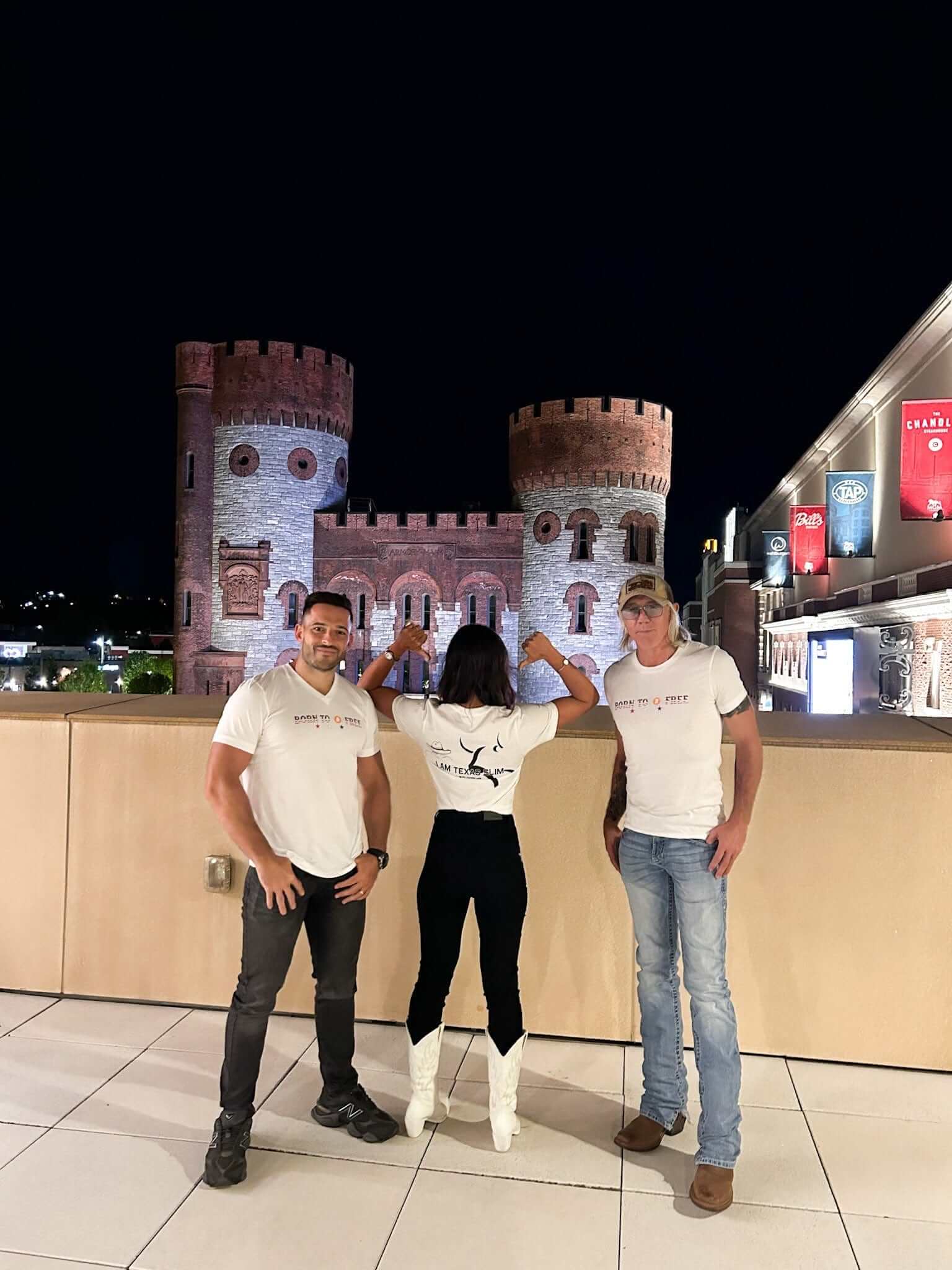 Three people wearing Feeding Forward T-shirts pose in front of a castle-like structure at night.