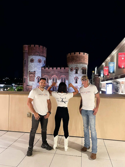 Three people wearing Feeding Forward T-shirts pose in front of a castle-like structure at night.