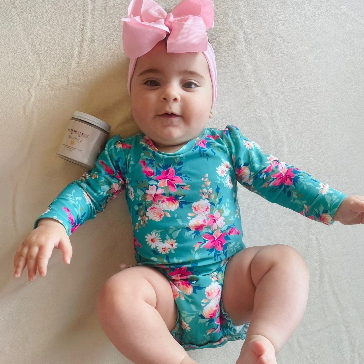 Happy baby wearing a floral swimsuit with a pink bow, next to a jar of Mineral Based Sun Armor Bundle.
