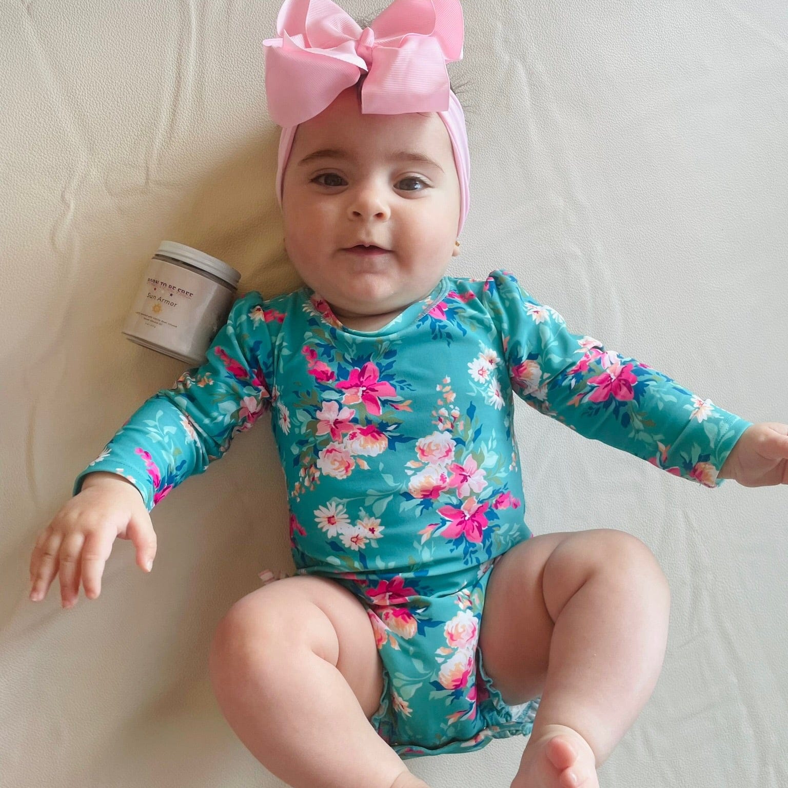 Happy baby wearing a floral swimsuit with a pink bow, next to a jar of Mineral Based Sun Armor Bundle.
