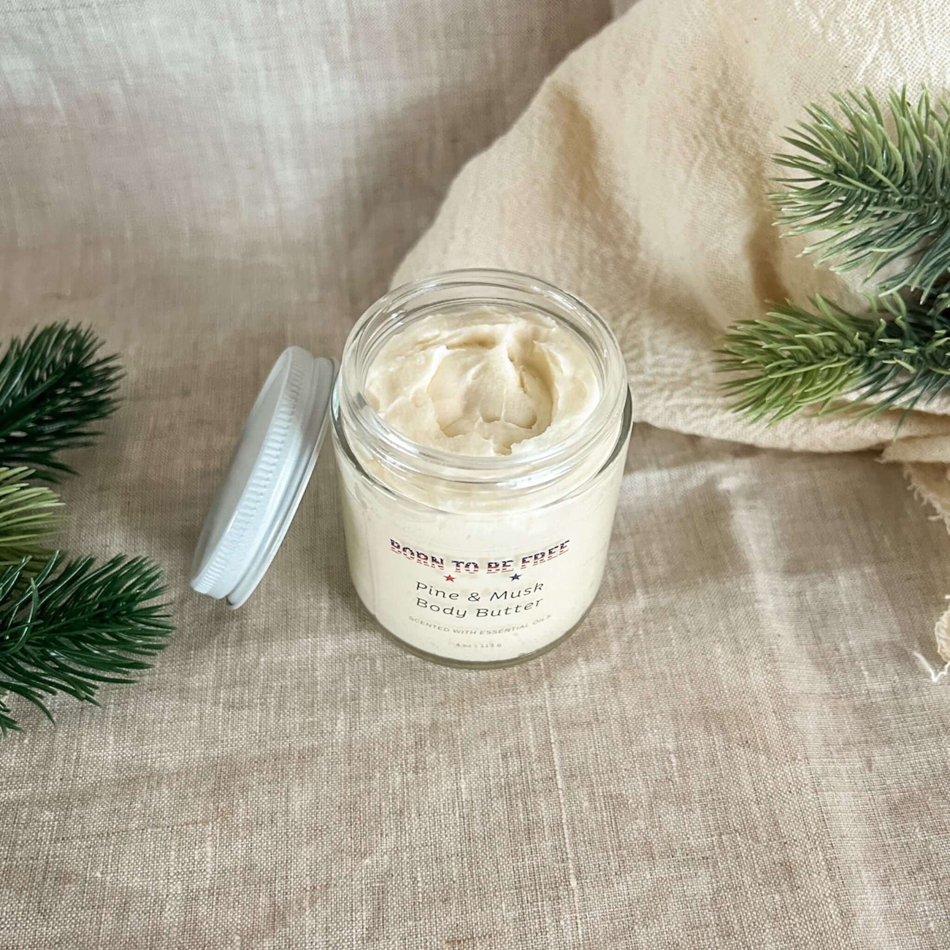 Pine & Musk Body Butter in a glass jar surrounded by natural greenery on a textured beige fabric background.