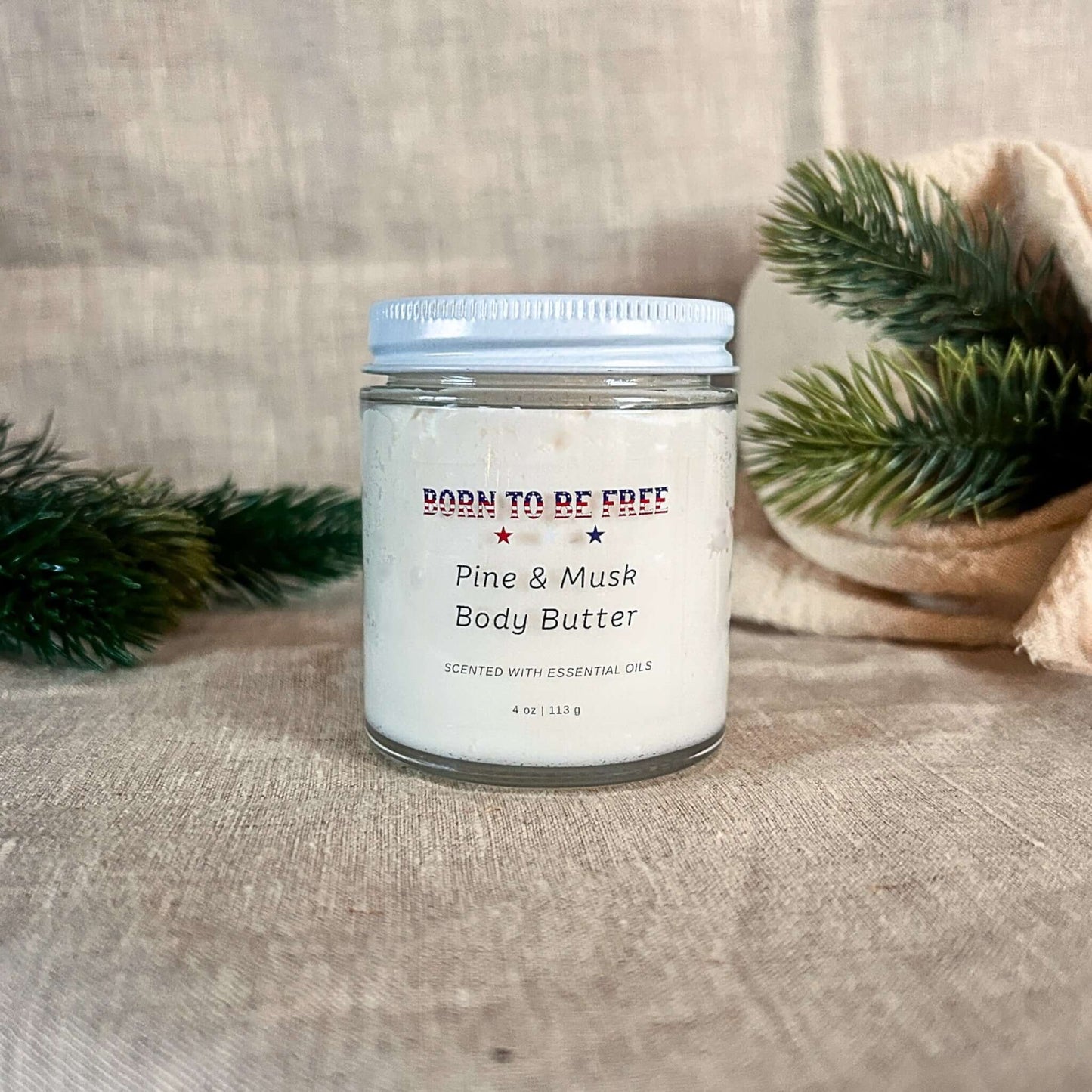 Pine & Musk Body Butter in a jar surrounded by pine branches, showcasing its soothing, natural essence.