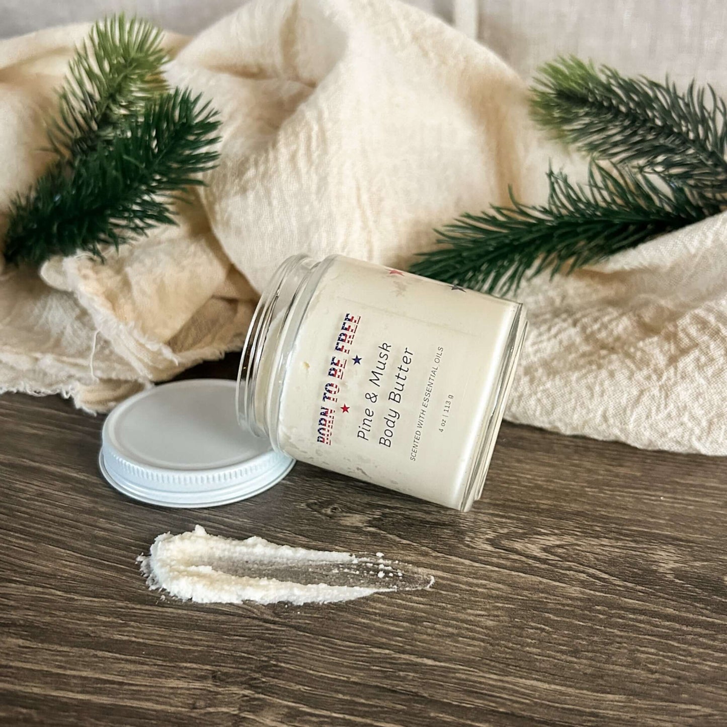 Pine & Musk Body Butter in a glass jar surrounded by natural elements like fabric and pine leaves.