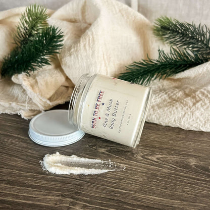 Pine & Musk Body Butter in a glass jar surrounded by natural elements like fabric and pine leaves.