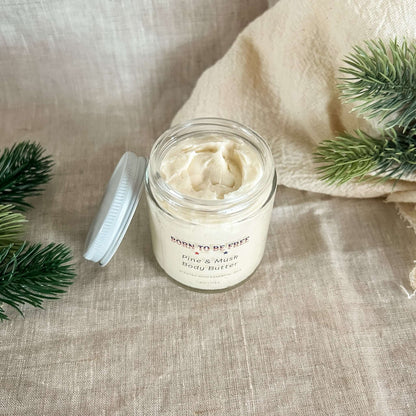 Pine & Musk Body Butter in a glass jar surrounded by natural greenery on a textured beige fabric background.