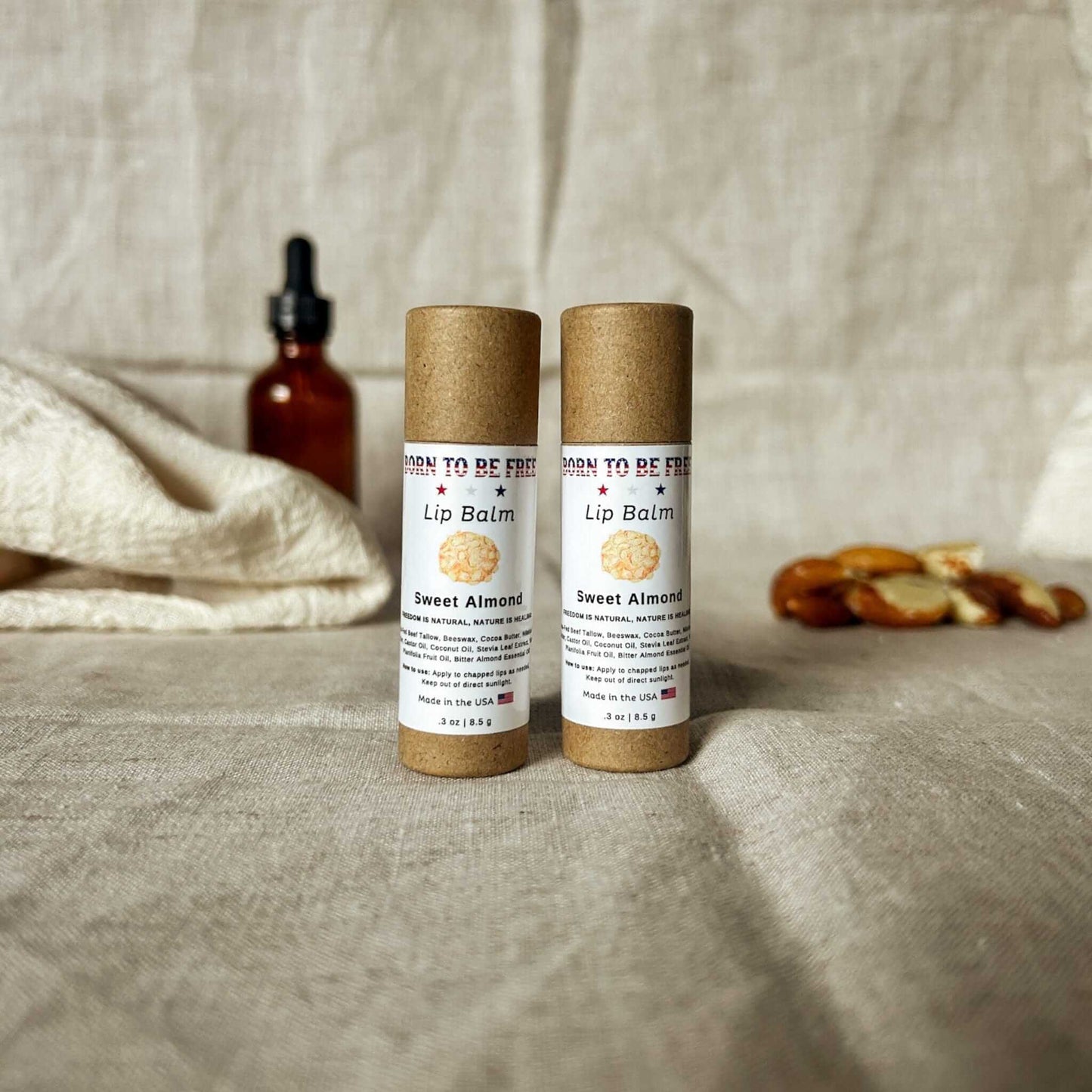 Two Sweet Almond Lip Balm tubes on a textured fabric background with almonds and a dropper bottle.
