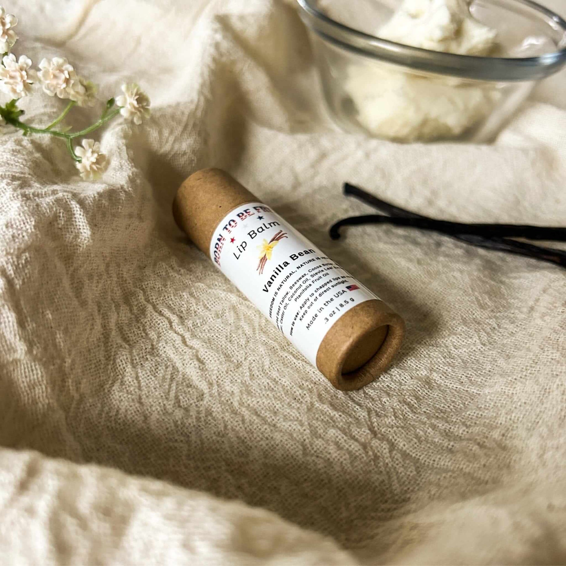 Vanilla Bean Lip Balm in eco-friendly packaging with flowers and vanilla pods in the background, promoting natural lip care.